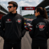 Black racing jacket with lion logo front and back worn by man and woman on racetrack with race car in background motorsport style Title: